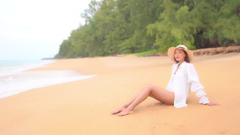A-pretty-woman-in-a-white-cover-up,-and-sun-hat-sitting-on-a-sandy-tropical-beach