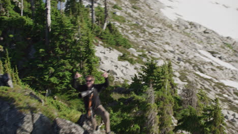 Photographer-triumphantly-raises-both-hands-and-throws-peace-sign-and-shaka-on-mountain-ridge,-Tim-Durkan-wide-helicopter-fly-over-SLOW-MOTION