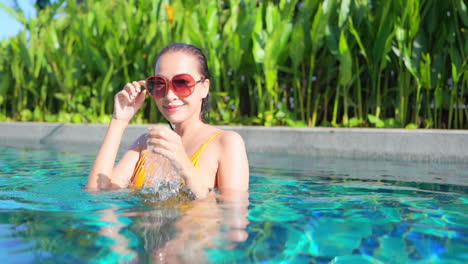 An-attractive-woman,-chest-high-in-a-swimming-pool,-looks-at-the-camera-as-she-adjusts-her-sunglasses