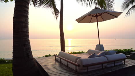 A-couchioned-couch-wedged-between-palm-tree-trunks-sits-empty-as-the-sun-sets-over-a-pink-ocean-horizon