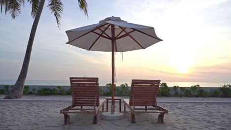 With-the-sun-setting-in-the-distance,-an-empty-pair-of-beach-loungers-sit-under-a-sun-umbrella-on-the-beach