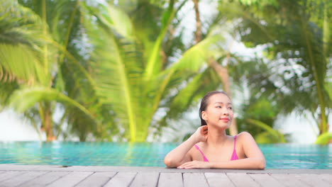 An-attractive-young-woman-in-a-swimming-pool-leans-along-the-edge-of-the-pool-as-she-looks-off-to-her-left-and-right,-palm-trees-in-the-background