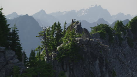Camera-cirlces-right-past-rocks-and-trees-to-reveal-photographer-on-mountain-ridge,-Tim-Durkan-wide-tracking-helicopter-counterclockwise-orbit-SLOW-MOTION
