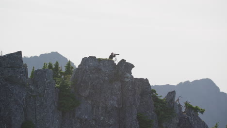 Photographer-kneels-on-mountain-ridge-as-silhouette-against-hazy-sky,-Tim-Durkan-wide-tracking-helicopter-counterclockwise-orbit-SLOW-MOTION