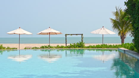 A-slight-ocean-breeze-ruffles-the-palm-fronds,-water-in-a-resort-swimming-pool,-and-beach-umbrellas-against-an-ocean-horizon