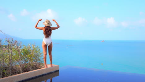 Back-to-the-camera-A-pretty-young-woman-standing-on-the-edge-of-a-resort-pool-looks-out-at-the-ocean-view-and-rejoices