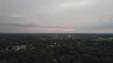 Aerial-drone-forward-moving-shot-over-an-old-mill-alongside-a-small-town-surrounded-by-dense-vegetation-on-a-cloudy-sunset