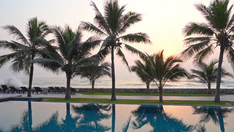 Pan-over-a-resort-swimming-pool-lined-with-palm-trees,-ocean-waves-roll-in-as-the-sunsets-in-the-background