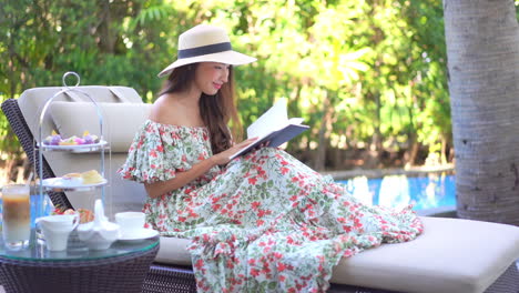 A-pretty-young-woman-in-a-sundress-and-straw-sun-hat-sits-on-a-chase-lounge-as-she-reads-her-book