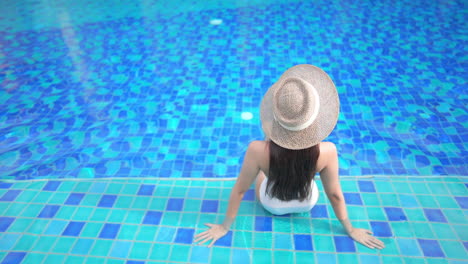 Back-to-camera,-A-woman-is-sitting-on-the-shallow-step-of-a-swimming-pool