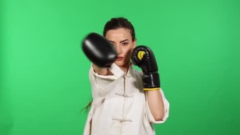 Junge-Brünette-Frau-Mit-Boxhandschuhen-Und-Weißem-Kung-Fu-Wushu-Unifrom-Stanzen-In-Richtung-Kamera-Mit-Grünem-Bildschirm-Im-Hintergrund