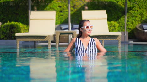 While-standing-in-waist-high-pool-water-an-attractive-young-woman-adjusts-her-sunglasses