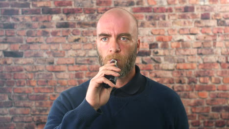 Portrait-of-a-man-vaping-indoors-in-front-of-a-brick-wall-looking-directly-at-the-camera-with-cloud-of-smoke