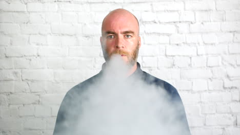 Portrait-of-a-man-vaping-indoors-in-front-of-a-white-wall-looking-directly-at-the-camera-with-cloud-of-smoke
