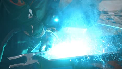 Worker-in-welder's-mask-welding-metal-parts-with-blue-high-temperature-sparkles---slow-motion