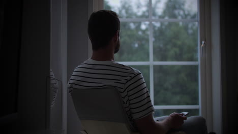 Male-adult-sitting-in-front-of-window-in-dark-room-on-overcast-rainy-day