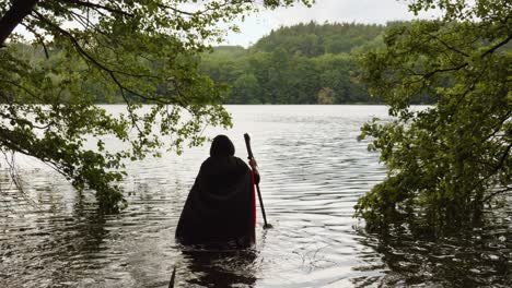 Back-View-Of-Grim-Reaper-In-Black-Hooded-Robe-Walking-Into-The-Water-With-Scythe