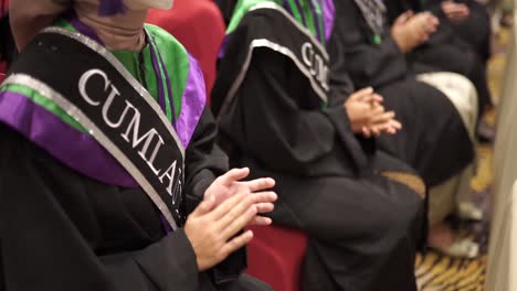 Close-Up-Happy-graduates-in-hats-and-robes-siting-in-row-and-applauding