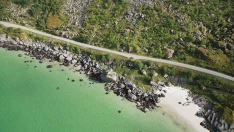 Aerial-shot-showing-the-crystal-clear,-bright-green-sea,-surrounded-by-a-scenic-road-in-Norway,-Lofoten-with-a-black-car-driving-on-it