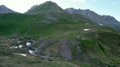 Aerial-drone-footage-slowly-approaching-a-Swiss-Alpine-hut-set-amoungst-the-grassy-slopes-in-a-mountain-landscape-with-residual-patches-of-snow-and-alpine-meadows-in-Switzerland