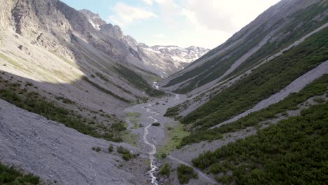 Aerial-drone-footage-flying-down-and-through-a-dramatic-glacial-valley-surrounded-by-a-steep-mountains-and-pine-trees-with-patches-of-snow-and-an-alpine-river-in-Switzerland