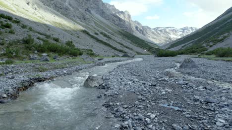 Aerial-drone-footage-flying-low-and-close-to-a-fast-flowing-river-in-a-glacial-valley-surrounded-by-a-steep-and-dramatic-mountain-landscape-with-residual-patches-of-snow-in-Switzerland