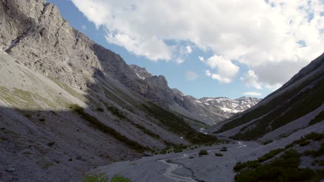Aerial-drone-footage-slowly-descending-into-a-glacial-valley-surrounded-by-a-steep-and-dramatic-mountain-landscape-with-residual-patches-of-snow-and-an-alpine-river-in-Switzerland