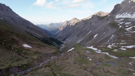 Aerial-drone-footage-facing-down-a-glacial-valley-and-slowly-reverseing-through-a-dramatic,-jagged-mountain-landscape-with-residual-patches-of-snow-and-alpine-meadows-in-Switzerland