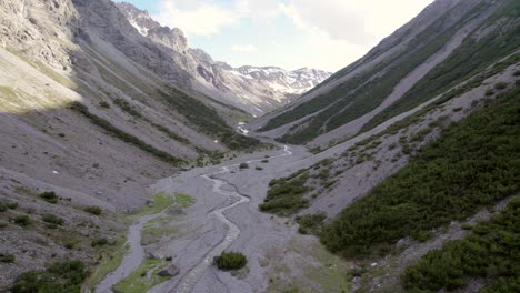 Imágenes-Aéreas-De-Drones-Que-Descienden-Lentamente-Hacia-Un-Espectacular-Valle-Glacial-Rodeado-De-Montañas-Empinadas-Y-Pinos-Con-Parches-De-Nieve-Y-Un-Río-Alpino-En-Suiza