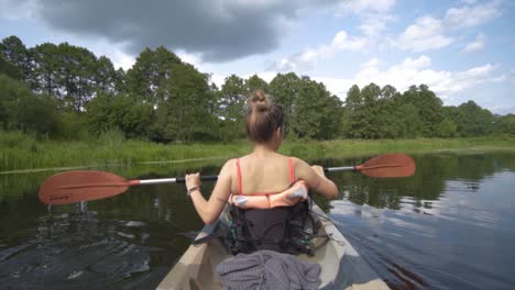 Blonde-girl-with-her-back-to-the-camera-paddling-a-kayak-in-slow-motion-on-a-river-surrounded-by-forest