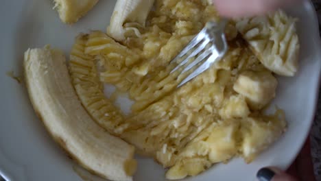Overhead-close-up-of-hand-mushing-banana-with-fork-in-white-bowl