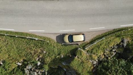 Slowly-approaching-aerial-shot-showing-a-golden-car-parked-on-the-side-of-the-road-surrounded-by-grass-and-trees-and-rocks