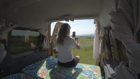 Blonde-digital-nomad-girl-taking-pictures-with-phone-of-the-landscape-from-the-back-of-a-camper-van