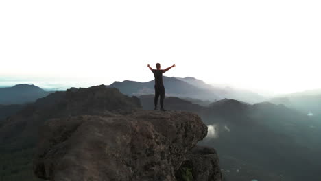 Mann-Auf-Dem-Roque-Nublo-In-Gran-Canaria-Streckt-Seine-Arme-Aus-Und-Genießt-Die-Aussicht,-Während-Die-Drohne-Im-Sonnenuntergang-Einen-Orbitflug-Macht,-Während-Nebel-über-Den-Felsen-Zwischen-Den-Wäldern-Liegt