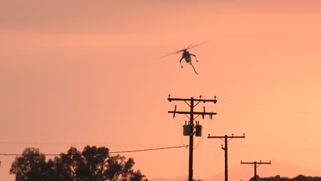 Following-a-firefighter-helicopter-flying-over-power-lines,-during-a-hazy-sunset