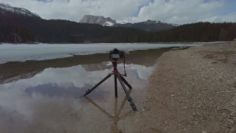 A-camera-on-a-tripod-sitting-in-lake-Crno-Jezero,-Albania-taking-landscape-photos-of-snow-capped-mountains