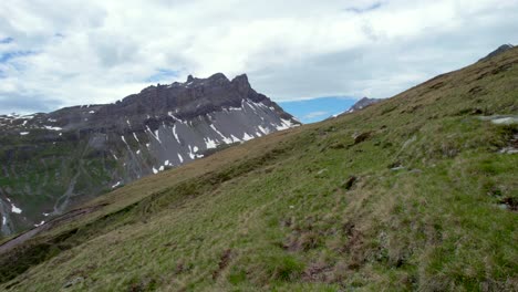 Aerial-drone-footage-flying-slow-and-low-over-a-hiking-trail-leading-along-grassy-slopes-towards-a-dramatic,-jagged-mountain-ridge-with-residual-patches-of-snow