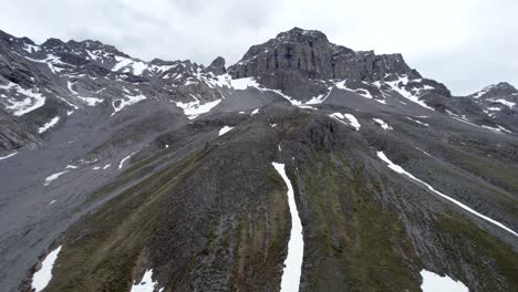 Aerial-drone-footage-flying-slowly-upwards-through-a-dramatic,-jagged-mountain-landscape,-looking-towards-an-imposing-grey-rocky-mountain-peak-with-residual-patches-of-snow