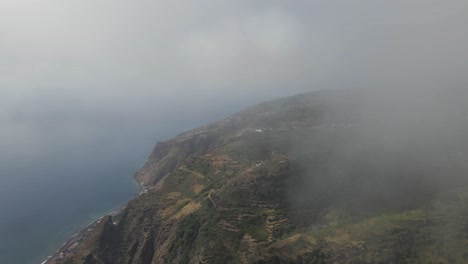 Aerial-view-of-the-Calheta-parish-in-the-Madeira-Island,-some-mist-clearing-from-the-camera-showing-the-green-mountains-and-the-blue-ocean