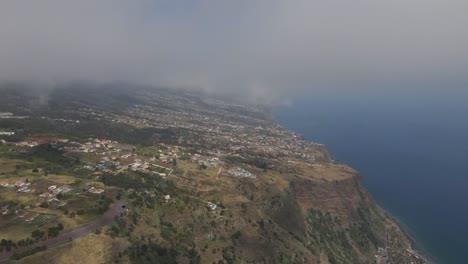 Aerial-view-of-the-Calheta-parish-in-the-Madeira-Island,-flying-above-the-houses-and-under-the-clouds-with-the-blue-ocean-on-the-right-side-of-the-camera
