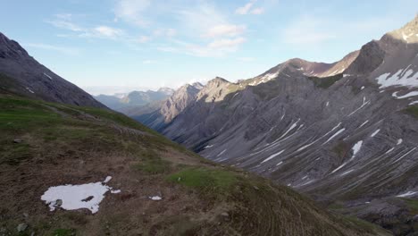 Aerial-drone-footage-slowly-rising-upwards-and-looking-across-a-dramatic,-jagged-mountain-landscape-with-residual-patches-of-snow-and-alpine-meadows-looking-down-a-glacial-valley-in-Switzerland