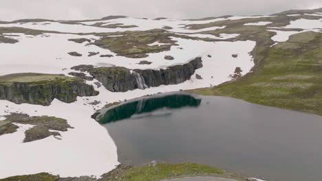 Vista-Desde-La-Carretera-De-Montaña-De-La-Impresionante-Cascada-Que-Cae-Sobre-Un-Acantilado-Rocoso-Cubierto-De-Nieve-En-Aurland,-Noruega