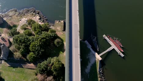 Cars-driving-over-the-bridge-over-the-Ulla-River-with-Viking-boats-on-the-jetty-below,-sunny-afternoon,-drone-shooting-traveling-forward-zenith
