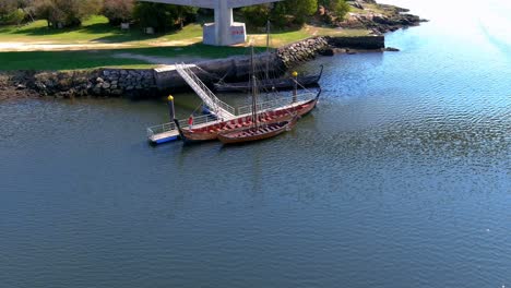 Two-people-observe-and-browse-on-the-jetty-some-Viking-ships-restored-imitation-of-the-originals-in-the-Ulla-River,-sunny-summer-afternoon,-drone-shot-orbits-zenithal
