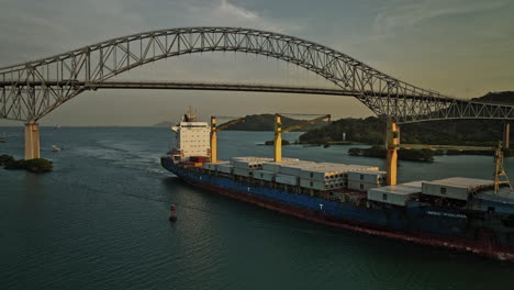 Panama-City-Aerial-v51-cinematic-low-level-fly-around-commercial-cargo-ship-at-puerto-balboa-capturing-bridge-of-americas-with-glowing-sun-at-sunset-golden-hours---Shot-with-Mavic-3-Cine---March-2022