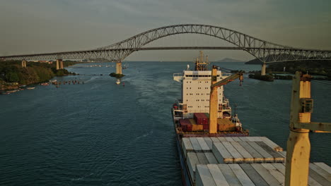 Panama-City-Aerial-v53-cinematic-low-level-drone-flyby-a-bulk-carrier-cargo-ship-around-puerto-balboa-harbor,-fly-under-bridge-of-americas-with-sunset-views---Shot-with-Mavic-3-Cine---March-2022