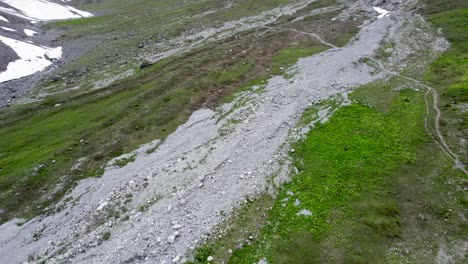 Aerial-drone-footage-tilting-slowly-up-to-reveal-an-alpine-meadow-and-grassy-slope-set-amongst-a-rugged,-glacially-carved-mountain-landscape-with-patches-of-snow-and-a-hiking-trail-in-Switzerland