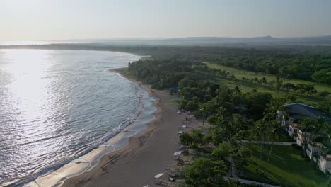 Cinematic-drone-shot-over-Playa-Dorada-Beach-with-sun-reflection-on-water-surface-and-forest-landscape-at-sunset---Puerto-Plata,Dominican-Republic