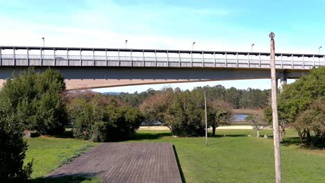 Autobrücke-über-Die-Ulla-Gebaut-Hinderlich-Inmitten-Eines-Gartens-Mit-Bäumen-Und-Sträuchern-Am-Wasser,-Ein-Sonniger-Nachmittag,-Drohne-Schießen-Vorwärts