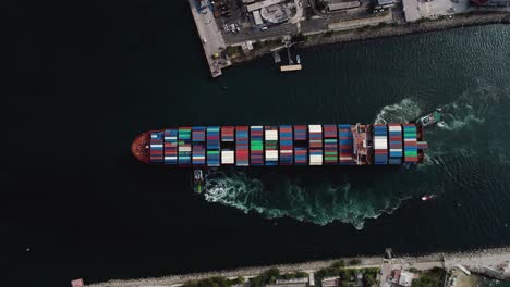 Mega-large-ULCV-container-ship-fully-loaded-with-crates,-sailing-away-from-the-harbor---Birds-eye,-aerial-view